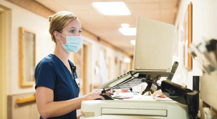 Registered nurse wearing a face mask documents medicine provided to a patient into a laptop computer at the nursing station at work, Indiana, USA