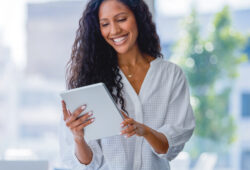 Young business woman using a digital tablet. She is casually dressed and smiling with a window behind her. She is sitting on a board room table with a laptop computer on it