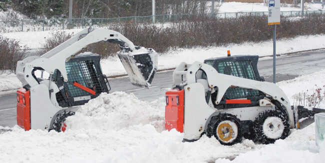 Two multi purpose mini front end loaders remove snow from a bus stop lane and sidewalk.