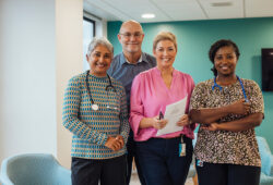 A portrait of four healthcare professionals standing in a common room in a hospital in Newcastle upon Tyne, North East England. They are standing side by side, looking at the camera and smiling.
