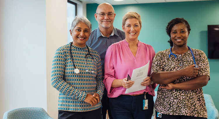 A portrait of four healthcare professionals standing in a common room in a hospital in Newcastle upon Tyne, North East England. They are standing side by side, looking at the camera and smiling.